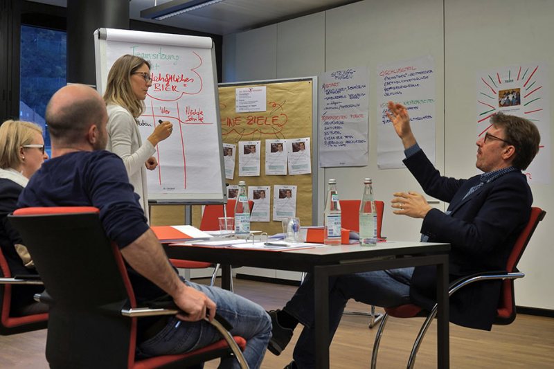 Sitzungssimulator-Gruppe-Sitzungsleiterin-Tisch-stehen-Flipchart-E9736b