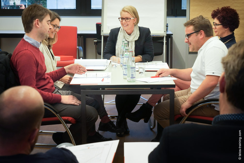 Sitzungssimulator Gruppe Sitzungsleiterin Tisch lächeln Beobachter Zuschauer / Foto: TELOS - E9706bdoF